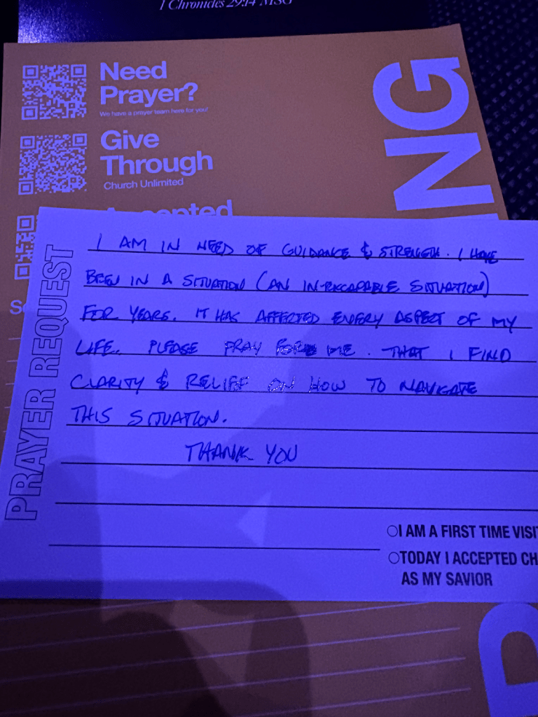 A handwritten prayer request card expressing a need for guidance and strength in a challenging situation, placed on a table with church-related materials.